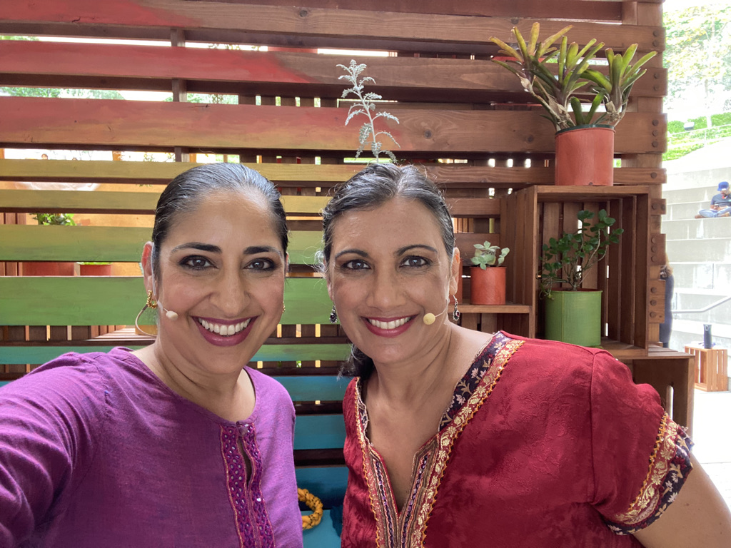 Sheetal Gandhi and Shyamala Moorty close up with lavalier mics, smiling before going on stage.  