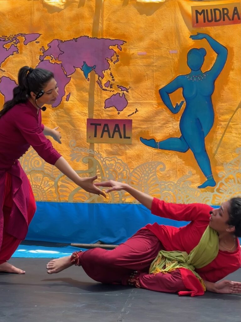 One female dancer with long hair, reaching down to help another female dancer with short hair lying on her side. their hands are almost meeting as they reach to each other. Both are wearing red in front of a yellow background.