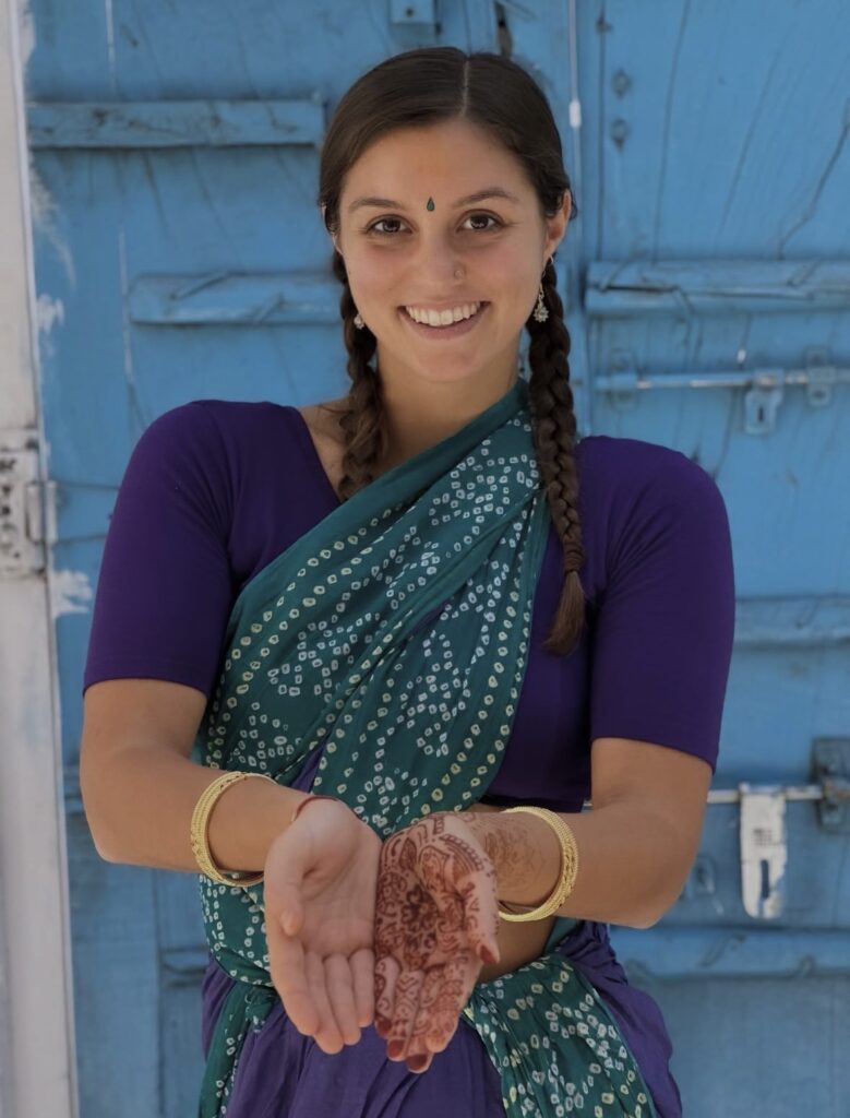 Tulsi Shah is smiling warmly with her hands open to the viewer with decorative henna on one palm.  She is wearing an Indian dance outfit with her hair in two long side braids.  